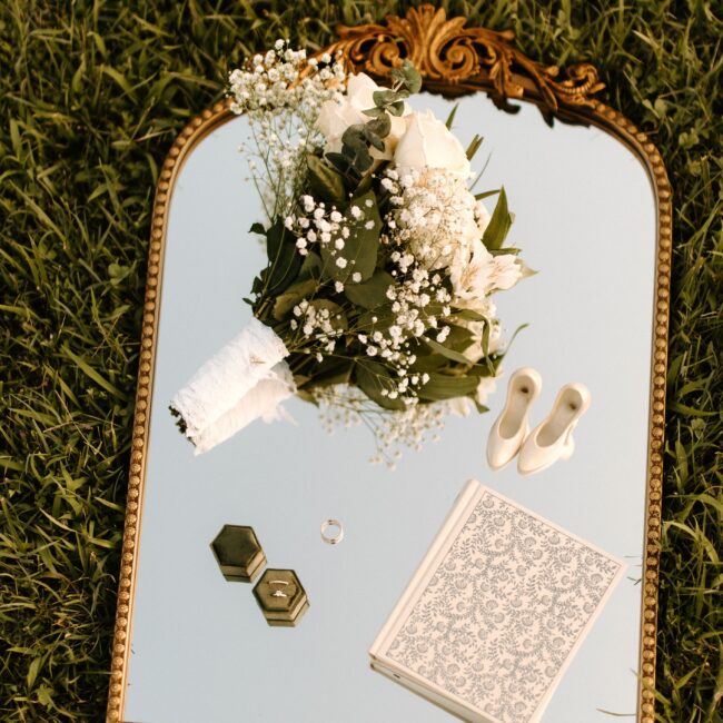 Elegantes Hochzeits-Flatlay, Ringe, Brautstrauß und Papeterie auf einem Spiegel mit goldenem Rand